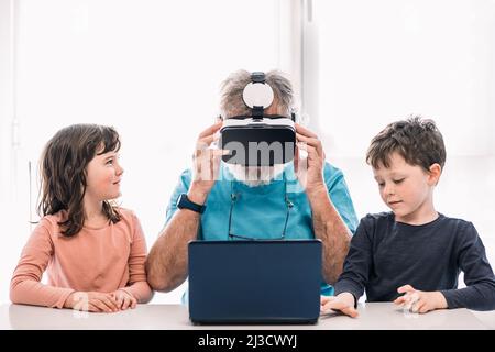 Bearded grandfather in modern VR headset playing videogame on laptop while sitting at table near cute children in light room Stock Photo