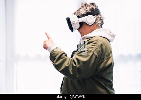 Side view of female wearing outerwear in modern white VR goggles experiencing virtual reality while pointing and standing on street of city Stock Photo