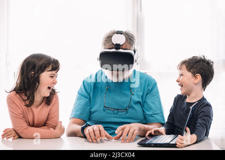 Bearded grandfather in modern VR headset playing videogame on laptop while sitting at table near cute children in light room Stock Photo