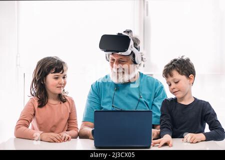 Bearded grandfather in modern VR headset playing videogame on laptop while sitting at table near cute children in light room Stock Photo