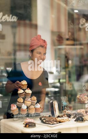 Hispanic Woman in headscarf arranging desserts on table near window while working in bakery in daytime Stock Photo