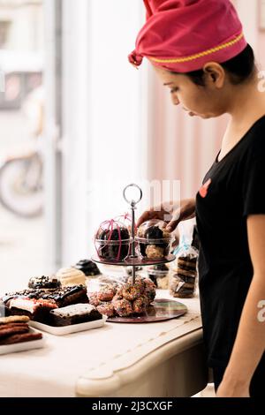 Hispanic Woman in headscarf arranging desserts on table near window while working in bakery in daytime Stock Photo