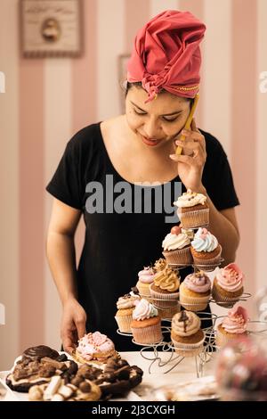 Hispanic Woman arranging pastry on table and talking on cellphone during work in bakehouse in daytime Stock Photo