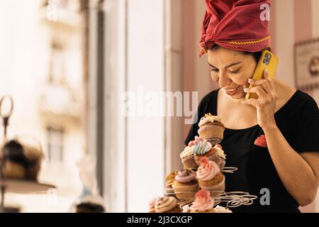 Hispanic Woman arranging pastry on table and talking on cellphone during work in bakehouse in daytime Stock Photo