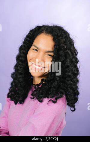 Positive African American female with black curly hair and looking at camera while standing in light studio on purple background Stock Photo