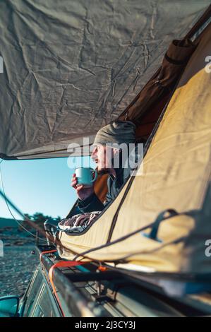 Male hiker in warm clothes lying on blanket in camping tent and drinking cup of coffee in morning and admiring nature at dawn Stock Photo