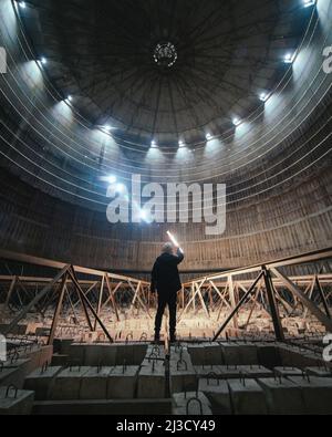 Unrecognizable person with bright lamp standing on concrete blocks and examining large dome inside desolate power plant Stock Photo