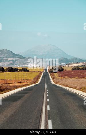 Empty straight asphalt roadway running through fields to in countryside to high mountains under blue sky Stock Photo