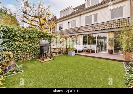Cozy wooden table and soft sofa and grill placed in backyard with green garden near contemporary residential building Stock Photo