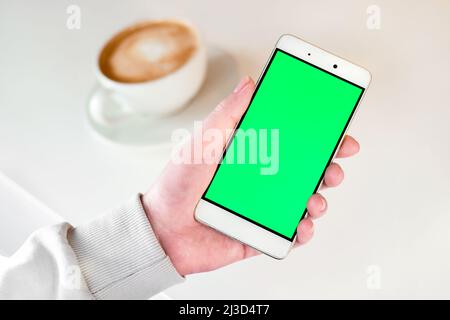 Hand holding green Screen Phone with a Cup of Cappuccino on white table. Woman using Mobile Phone with Chroma Key. Over shoulder View. Stock Photo