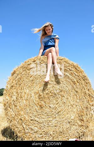 Young attractive blonde woman of white race sits barefoot on a round haystack and smiles Stock Photo