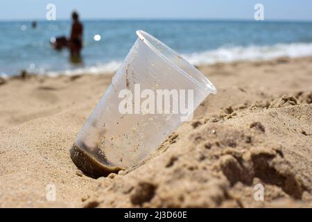 On the sandy shore of the sea beach lies a dirty plastic glass close-up. The background is very blurry Stock Photo