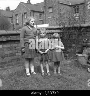 1961, historical, three girls, sisters, standing for a photo in the walled back garden of a victorian terraced house, Stockport, Manchester, England, UK. A metal bucket of the era, with lid can be seen. Stock Photo