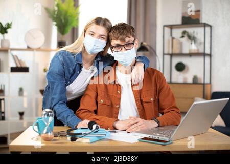 Students, man and woman in casual outfit and face masks posing on camera, sitting at the table and using laptop. Young partners cooperating or working remotely online from home Stock Photo