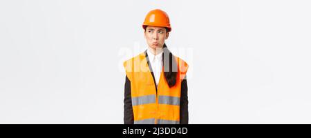 Gloomy and offended young asian female construction manager, industrial woman in safety helmet complaining or whining, pouting sad and looking upper Stock Photo