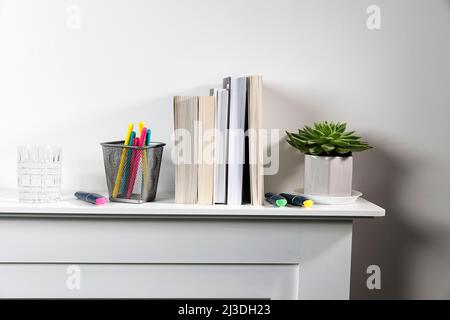 Echeveria in geometric pot, stack of books, pens in a pencil holder, felt-tip pens, are on shelf. Place for text Stock Photo