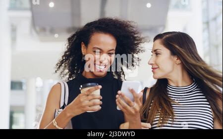 They are the best of colleagues. Cropped shot of two young businesswomen chatting and using a smartphone while walking in an office on a coffee break. Stock Photo
