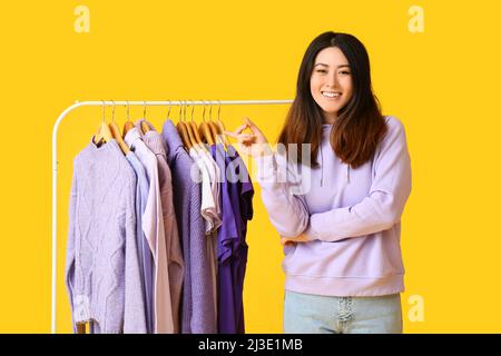 Young Asian woman pointing at rack with clothes in purple shades on yellow background Stock Photo