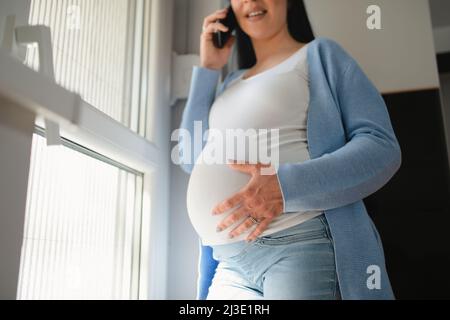 A pregnant black-haired caucasian holds a phone in her right hand and talks and touches her belly with her right hand. A woman stands next to the door. Stock Photo