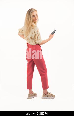 beautiful European blue-eyed blonde girl standing on her back and looking into the camera wearing shirt and pink trousers and sneakers holding a phone studio shot white background full shot copy space. High quality photo Stock Photo