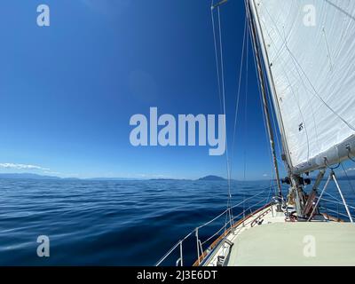 View of sailboat sailing along with sails up and tilted by the wind. Blue skies and mountains in the foreground, Strait of Georgia, Canada Stock Photo