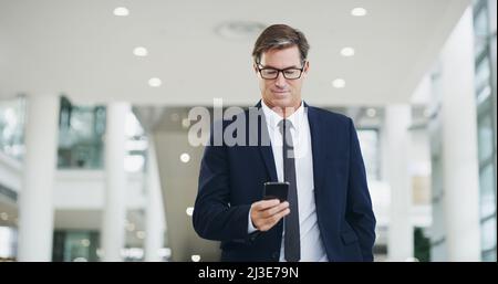 Time to connect with some colleagues. Cropped shot of a mature businessman using a smartphone while walking through a modern office. Stock Photo