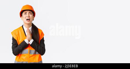 Desperate and reckless asian female construction manager, architect pleading god, wearing safety helmet, holding hands in pray, supplicating, asking Stock Photo