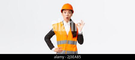 Imressed and surprised asian female construction manager pleased with amazing quality, standing in safety helmet and reflective jacket, showing okay Stock Photo