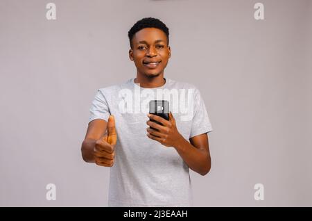 cute guy isolated over grey background smiling as he holds his phone and did thumbs up Stock Photo