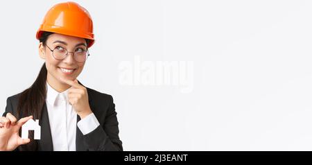 Creative smiling asian female architect thinking about new design during construction works, looking thoughtful with smile as holding small house Stock Photo