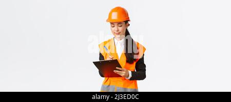 Pleased asian female construction engineer, architect taking notes in clipboard, writing down something during inspection at building area, wearing Stock Photo