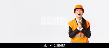 Desperate and reckless asian female construction manager, architect pleading god, wearing safety helmet, holding hands in pray, supplicating, asking Stock Photo