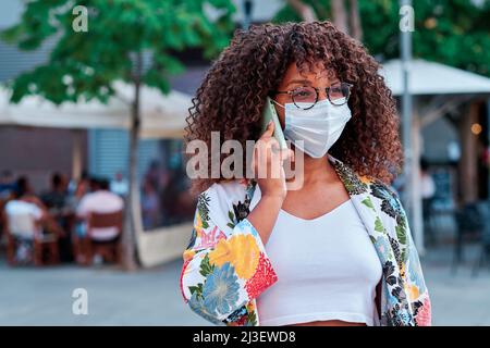 young afro american woman using mobile phone and wearing a face mask during coronavirus outbreak Stock Photo