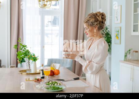 Girl cuts fruit salad with strawberries on cutting board with knife. Concept cooking sliced fresh fruits Beautiful woman with makeup and hairstyle in light dress enjoys morning and preparing breakfast Stock Photo