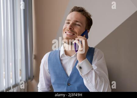 Pleasant handsome bearded man in shirt and waistcoat talking on phone, ordering taxi or food delivery to office at work. Smiling young caucasian businessman holding a call with partners clients. Stock Photo