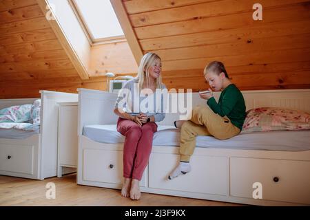 Happy boy with Down syndrome sitting on bed with his grandmother and using smartphone at home. Stock Photo