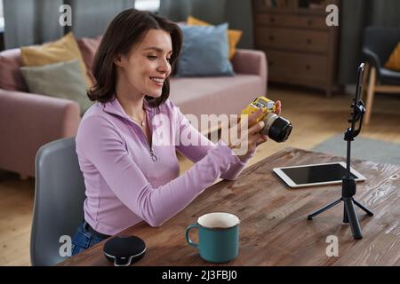 Positive attractive young Caucasian photographer sitting at table with smartphone on tripod and looking through cameraphotos Stock Photo