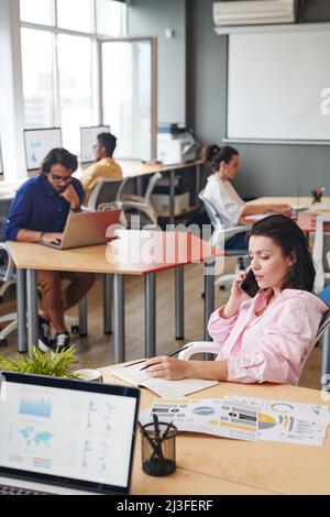 Serious young female analyst sitting at table and making notes in planner while talking to client by phone Stock Photo