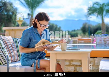 Middle aged woman resting near outdoor pool reading book Stock Photo
