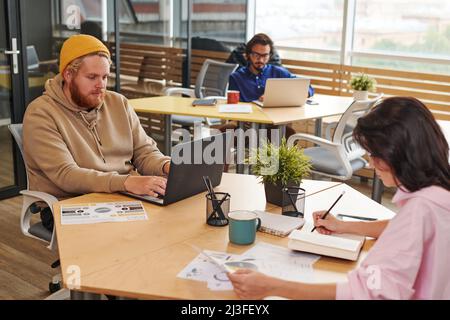Busy young managers sitting at tables in coworking space and working with laptops and papers Stock Photo