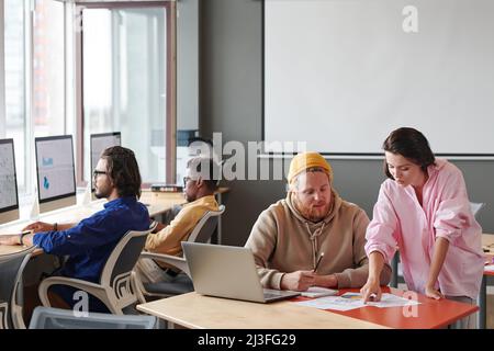 Young woman pointing at data in papers to colleague while they discussing project implementation in modern office Stock Photo