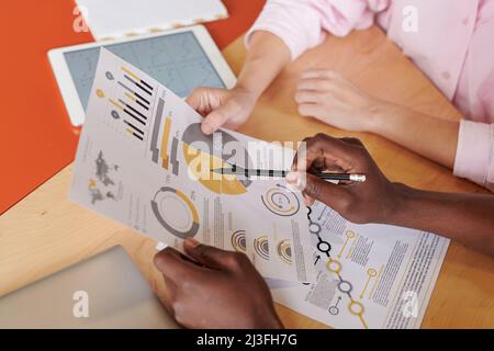 Close-up of unrecognizable colleagues sitting at table and holding paper while discussing statistical data together Stock Photo