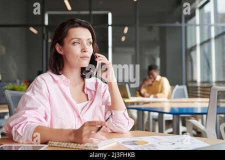 Pensive confident young business lady analyzing papers while talking by phone in coworking space Stock Photo