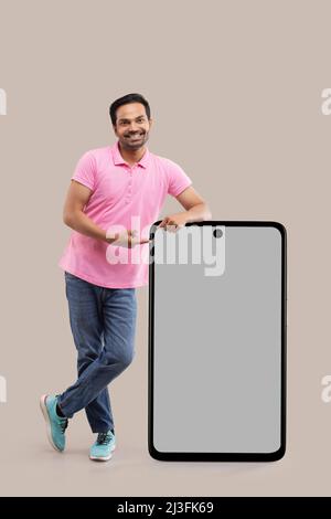 Smiling young man leaning on big smartphone and pointing with finger Stock Photo