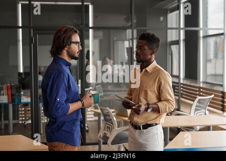 Young multi-ethnic managers standing in coworking space and discussing new project while one of them drinking coffee Stock Photo