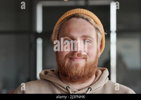 Headshot of satisfied young red-bearded Caucasian man in bright hat posing in modern office Stock Photo