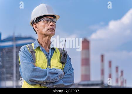 Senior engineer in his work cloths with white hard hat, safety glasses, and walkie talkie radio standing at the power plant site to supervise his proj Stock Photo