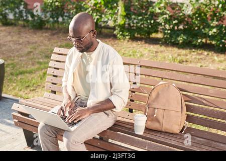 Busy young African-American man with bald head sitting on bench with satchel and coffee cup and using laptop outdoors Stock Photo