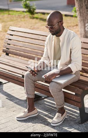 Serious young African-American entrepreneur in stylish casual outfit sitting on bench in city and working through smartphone Stock Photo