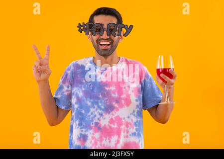 Funny young man in party eye glasses holding wine glass and gesturing with fingers